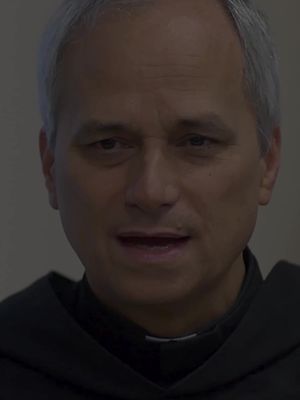 A medium shot of a man dressed in a black robe and white collar, looking directly at the viewer. The color palette is muted, with variations of gray and black dominating the image. The man's skin tone is light, and there's a slight shadow on his face, creating a somber atmosphere.