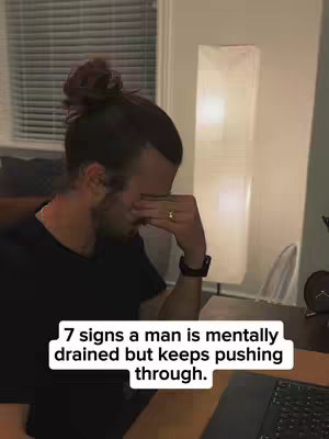A man with dark hair in a bun is shown from the side, covering his face with his hand. He appears to be in an office or living space, and text overlaid on a white background says, "7 signs a man is mentally drained but keeps pushing through."