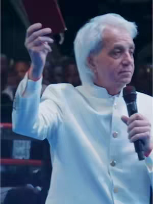 The image features a man with short, white hair, wearing a white suit and holding a microphone in one hand and a closed book in the other, likely addressing a crowd of people dimly visible in the background. The lighting appears to be from above.