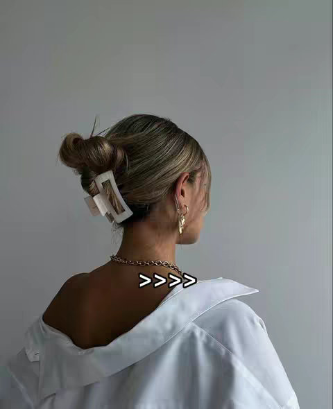 A woman with light brown, styled hair, is seen from behind, hair up in a messy bun with a large square hair clip. She is wearing a white button-down shirt with the collar down exposing her neck and back. A gold chain necklace is visible, as are gold hoop earrings. Arrows are overlaid on the image pointing towards the necklace.