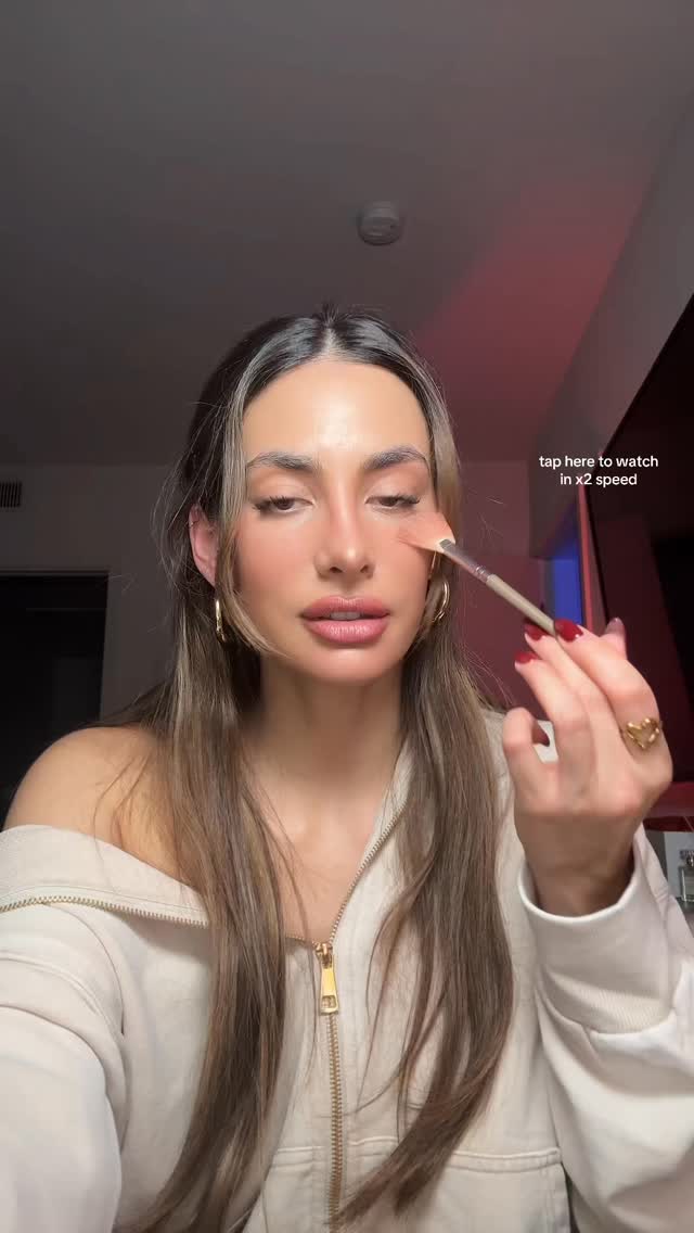 A woman is applying makeup, holding a brush to her cheek. Text overlay near the top right encourages watching in x2 speed.