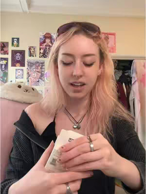 A young woman with light pink hair is centered in the frame, holding a small, unmarked package in both hands. She appears to be looking down, seemingly focused on the package, with a slight smile.