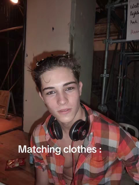 A young man with dark hair is the central focus, wearing a plaid shirt, headphones, and sunglasses on top of his head. The background is a dimly lit, cluttered area with scaffolding visible. Overlaid text reads "Matching clothes :".