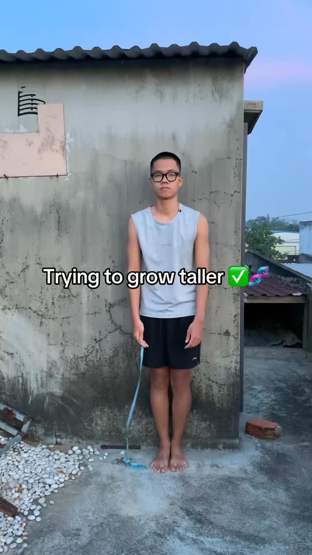 A young man, barefoot and wearing glasses, stands in front of a weathered concrete wall. He is wearing a sleeveless gray shirt and black shorts. The text overlay reads 'Trying to grow taller' with a checkmark.  The background shows a clear sky.
