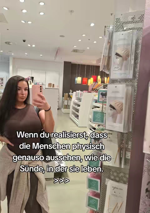 A young woman with long dark hair is taking a selfie in a store or retail environment. She is holding a pink smartphone in front of her face, positioned to capture both her and the surroundings. She wears a dark brown top, a light gray cardigan draped over her shoulders, and dark-colored pants. The background shows shelves with various retail items and display shelves. The text overlay adds a philosophical or contemplative note.