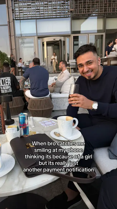 A man in a black shirt is seated at an outdoor table, smiling at his phone. He holds a glass of what appears to be a dark alcoholic drink and a watch on his left wrist. A cup of coffee rests on a saucer on the table, along with a used Red Bull can. Several other people are in the background, providing an outdoor setting.