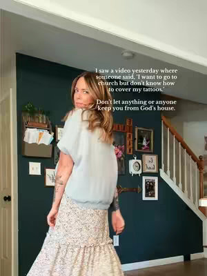 A woman in a light blue shirt and a patterned skirt poses in front of a dark teal wall adorned with various frames, a cross, and decorative shelves. The woman has tattoos visible on her arm and looks back at the camera with a gentle expression. The background includes a staircase and other home decor items, indicating an indoor domestic setting.