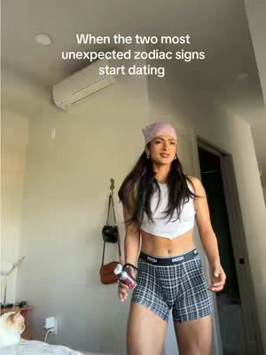 A woman in a white crop top, plaid boxer shorts, and a pink headscarf stands in a room. Text overlays the image, and the background features an air conditioner and a door. The woman appears to be holding a small container, likely a drink.