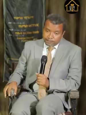 A man in a grey suit sits in a chair, holding a microphone. Behind him is a promotional banner with Amharic text. The upper right corner has a logo.