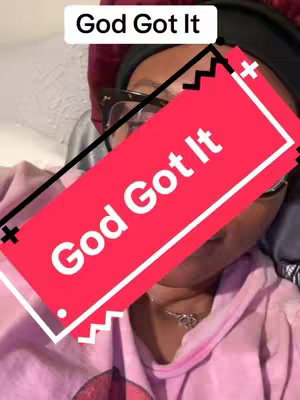 A selfie-style image with a person with dark hair and glasses. The person dons a black headband and is smiling while overlaid by two text elements. The person is wearing a pink hoodie and a delicate necklace. The top and the main text boxes emphasize the message 'God Got It' against a pink background and a white font. The photo is taken from the point of view of the content creator.