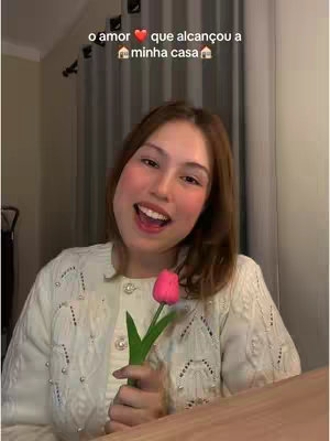 A young woman with light brown hair smiles and holds a pink tulip in front of a blurred background. The woman wears a white knit cardigan with pearl-like buttons. Text overlay above the woman reads "o amor que alcançou a minha casa."