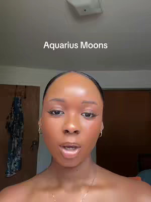 A close-up shot of a woman reveals her face, with the text 'Aquarius Moons' overlaid at the top. The woman has dark hair pulled back, and wears makeup with emphasis on her eyes and lips. The background is minimally visible, with a door and a ceiling light partially in view.