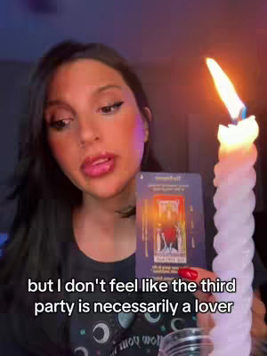 A young woman is holding a tarot card in front of her, discussing its meaning to the viewer. She has a close-up shot, with a lit candle providing warm light from the side. Her facial expression is focused, and the background is dimly lit.