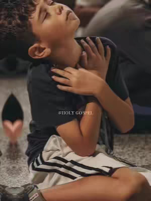 A young boy with light skin sits with his eyes closed and hands clasped at his chest, appearing in a moment of worship or prayer. He is angled slightly upwards, and the background is blurred. A small, dark heart-shaped image is to the left of the boy. The photo has muted colors and a soft focus.