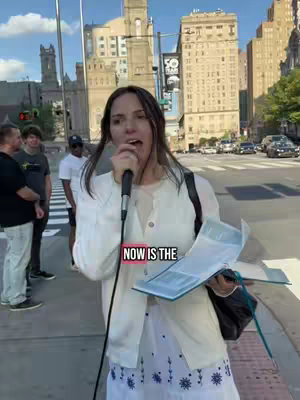 A woman with dark hair stands on a city street, holding a microphone and a book. She wears a white outfit with a floral-patterned skirt. The background includes buildings and pedestrians. Text overlayed on the image reads "NOW IS THE".