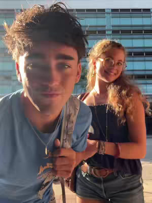 A selfie-style image featuring two young people standing outdoors. On the left, a young man with dark hair gazes at the camera; he is wearing a light blue t-shirt and a backpack strap. To the right, a young woman with blonde wavy hair smiles at the camera, wearing glasses and a necklace. They are both standing in front of a modern building.
