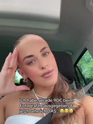A woman is seated in a car, framed from a slightly low angle with the camera angled up. The woman has fair skin, blonde hair pulled back, and blue eyes. She is wearing a white top and has a necklace. Her left hand rests on her head. Overlaid text near the bottom. Trees are visible outside the car window.