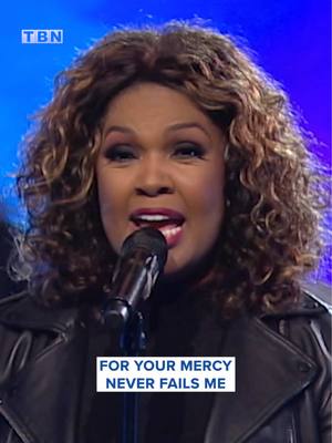 The image shows a woman with curly brown hair, wearing a black leather jacket and singing into a microphone. The background is a gradient of blue. A white box at the bottom of the screen contains the text: "FOR YOUR MERCY NEVER FAILS ME". The logo "TBN" is visible in the top left corner. The woman's expression is one of intense emotion, suggesting a performance of a gospel song.