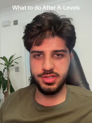 A close-up shot of a young man with dark hair and a beard, looking directly at the camera. The image is likely a selfie or vlog-style shot, framed to focus on his face. Behind him is a white wall and a faint glimpse of a leafy plant.