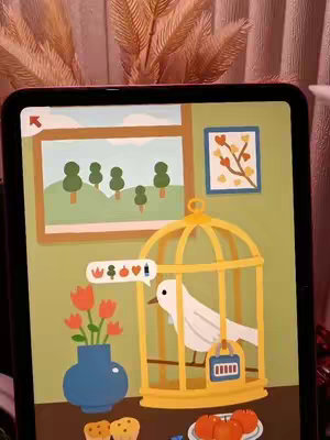 An iPad screen displays a whimsical game scene. The game shows a cartoon interior with a window looking out onto trees, a framed picture on the wall, a blue vase with orange tulips, a bird in a yellow cage, and various cartoon food items. The background behind the iPad appears to be a pinkish fabric. The scene has a warm, inviting aesthetic with soft colors.