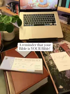 A photo of a work desk with a laptop, plants, notebooks, and a Bible. The laptop shows a video with a cartoon-like illustration. The scene is well-lit creating a cozy vibe