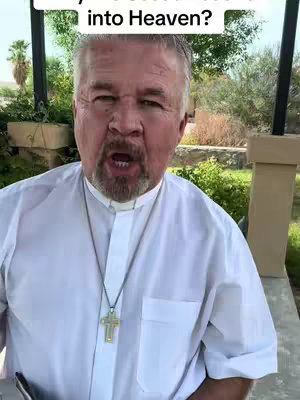 A man in a white clerical shirt and a cross necklace speaks directly to the camera outdoors. Green foliage and a building are visible in the background.