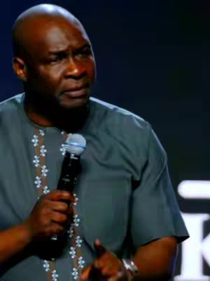 A medium shot shows a Black man addressing an audience, holding a microphone. He is wearing a patterned gray shirt. The background is dark, suggesting a stage or studio setting. The lighting is focused on the speaker.