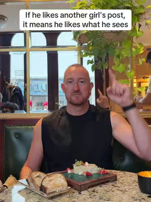 A man with a shaved head, wearing a black sleeveless shirt, is seated in a restaurant. The background is blurred, showcasing an indoor setting with wooden accents and green foliage. He points upwards with his right index finger, with food and some bread on the table in front of him. Above his head is a white text overlay.