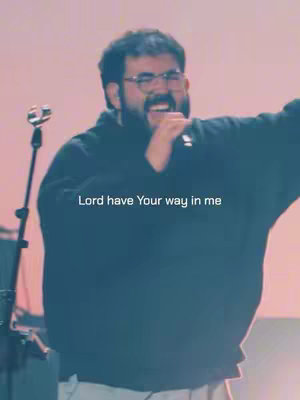 In a medium shot, a man with a beard, glasses, and a black sweater is on stage holding a microphone, singing into it. Underneath him, the caption reads, 'Lord have Your way in me'. The backdrop has a soft gradient of pink and yellow.
