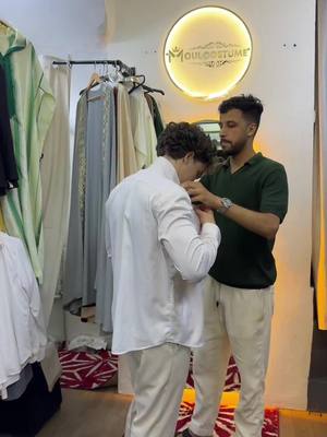 Two men are in a shop, one is dressed in a white shirt and beige pants being checked on his outfit by another man wearing a green polo shirt and beige pants. The shop interior is filled with clothing racks and a sign that reads 'MOULCOUSTUME' in a circular design.