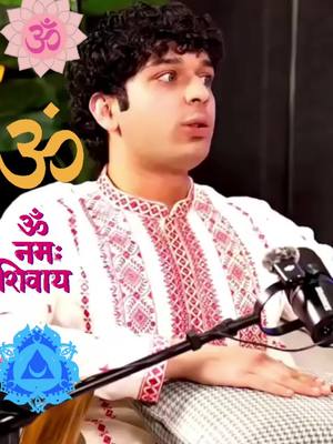 A man with curly dark hair is seated, looking slightly to his right with a contemplative expression. He wears a traditional white top with red patterned embroidery around the collar and front. A microphone is positioned to his right, and in the background, a backdrop displays the Om symbol with an image of a lotus flower. Further text in Hindi and a chakra icon are also visible, suggesting themes related to spirituality and meditation.