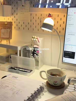 A well-lit desk setup featuring study materials and a cozy atmosphere. A white desk holds a notebook with handwritten notes, pens, and a cup of tea. A desk lamp provides warm light, enhancing the inviting feel of the space. The background includes a pegboard and an analog clock. The overall composition suggests study, peace, and productivity.
