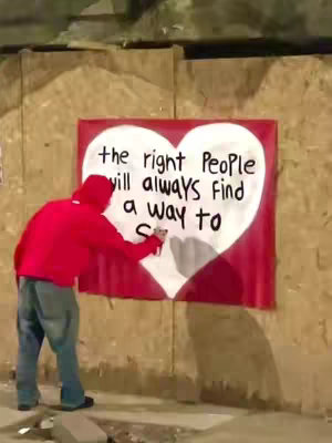 A person wearing a red hoodie and jeans is writing on a banner that is red with a white heart painted on it which has the words 'the right People will always Find a way to' written on it on a brown wall.
