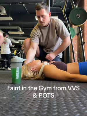 A woman lies on a gym floor, appearing unwell, with a man kneeling beside her. The scene is in a gym setting with equipment in the background and text overlayed at the bottom of the image.