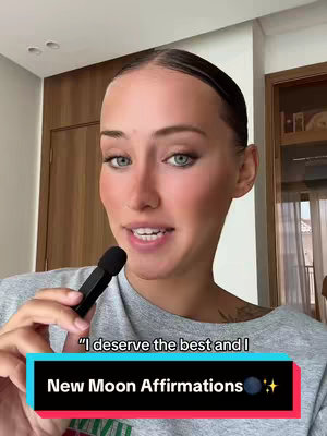 A woman with fair skin and light eyes is the primary focus, her head taking up most of the frame. She has her hair pulled back and is wearing a light colored t-shirt. Above her is a dark-colored rectangular bar with white text that reads "I deserve the best and I." Below, an additional bar displays the words "New Moon Affirmations" on a black background, with a small moon and star icons. She holds a microphone, and behind her is a room interior with neutral-toned walls and a wooden cabinet.