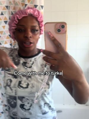 A woman in a pink shower cap and printed shirt is taking a selfie, pointing downwards with one hand while holding the phone in the other. The background features a bathroom with a patterned shower curtain. Text overlayed says "Come with me to turke".