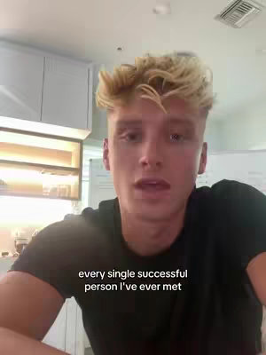 A young blonde man is centered in the frame, looking directly at the camera. He is wearing a black t-shirt, and his hair is styled with a tousled look. The background is a kitchen with white cabinets and a hint of a window. Text overlayed at the bottom reads 'every single successful person I've ever met'. The lighting appears bright and natural, suggesting an indoor setting during the day.