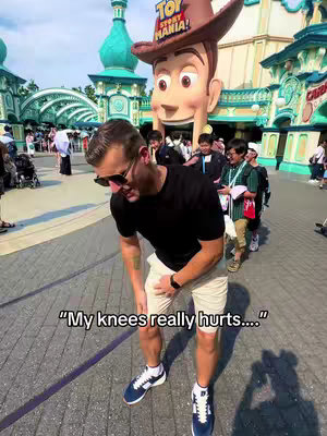A selfie taken in front of the Toy Story Mania entrance at Tokyo DisneySea during daytime. The main subject, a man wearing sunglasses, a black t-shirt, and khaki shorts, is looking down and holding his knee, with text overlaid along the bottom of the image. Behind him are several individuals walking in the same direction, and a giant Woody figure.