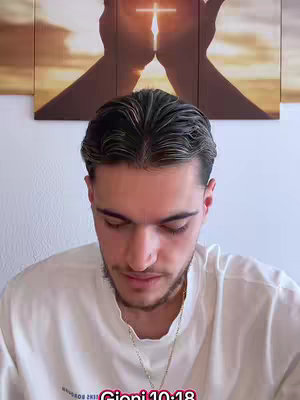 A young man is shown from the chest up, with his gaze directed downwards. He's wearing a white t-shirt and a gold chain necklace. The background includes a wall featuring a triptych art piece with a cross and hands, suggesting a religious theme, and the text "Gjoni 10:18" at the bottom.