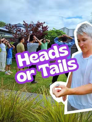 A group of people playing Heads or Tails in a backyard setting. A man in a white t-shirt is holding a coin and looking towards the viewers. The text overlay 'Heads or Tails' is displayed in a purple bubble-letter font with a white outline. Various people are in the background with their hands on their heads.