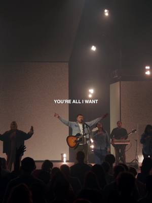 A live performance on stage with a band, a male singer in the center with arms outstretched, and text overlayed.