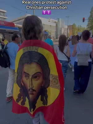 A person is walking away from the camera, wearing a red and gold cloth draped over their shoulders. The cloth depicts a portrait of Jesus. The background shows blurred figures walking toward a building in the distance.