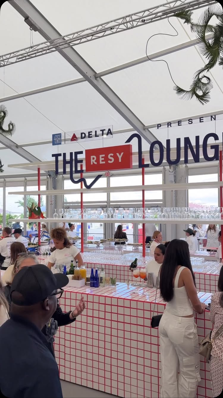 The cover image presents a lively outdoor lounge setting, likely for an event. The scene features a white tent structure with a partial view of the sky. Inside, a bar with a red and white checkered facade is active with several people. Behind the bar, there are shelves with glassware, and staff members are seen preparing drinks. Prominent signage displays "THE RESY LOUNGE," with logos of American Express and Delta Air Lines visible. The composition is a vertical shot, capturing a snapshot of the lounge environment with a focus on activity and branding.
