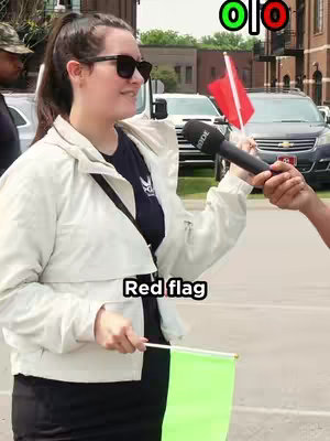 A woman wearing sunglasses and a white jacket, stands outdoors holding a green flag and is being interviewed. The words "Red flag" is overlaid in the center with the flag graphic colors (green and red) above her head.