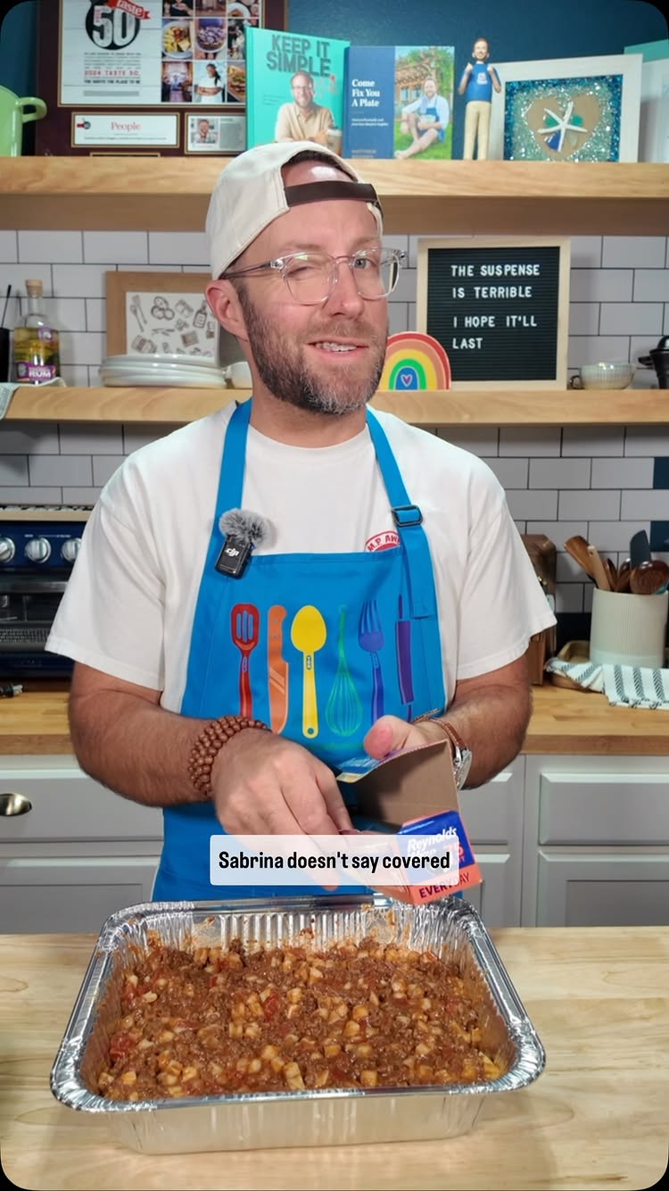 A man in a white t-shirt and blue apron winks while holding a container of casserole over a foil pan of food in front of a kitchen background. A text overlay reads, "Sabrina doesn't say covered."