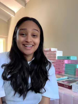 A woman with long, dark hair smiles at the camera, with a few products stacked behind her and a bright room behind them.