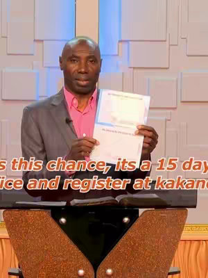A man in a gray suit and pink shirt stands behind a lectern. He holds a document in his hands, and text is overlaid near the bottom of the image.