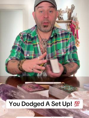 A man with short hair, wearing a grey cap, green and blue plaid shirt, and multiple jewelry, is holding a deck of cards in front of a table. In the foreground, there are tarot cards and crystals. Text overlay reads 'You Dodged A Set Up! 💯'.