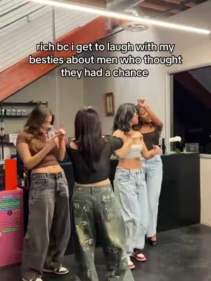 A group of four young women are captured in a casual shot. They are standing in what appears to be an indoor setting, possibly a store or salon, with simple lighting. The women are dressed in a variety of outfits, featuring crop tops and loose-fitting jeans. The color palette is fairly neutral, with denim blues and earth tones setting the clothing context. One woman's back is toward the camera, the others are smiling and pointing, and gesturing while facing each other. The hook text is at the top.
