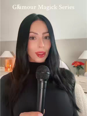 A woman with long, black hair is holding a microphone in front of a cream-colored background. The top of the image has the text "Glamour Magick Series". She is wearing a black turtleneck and has red lipstick with a light skin tone.